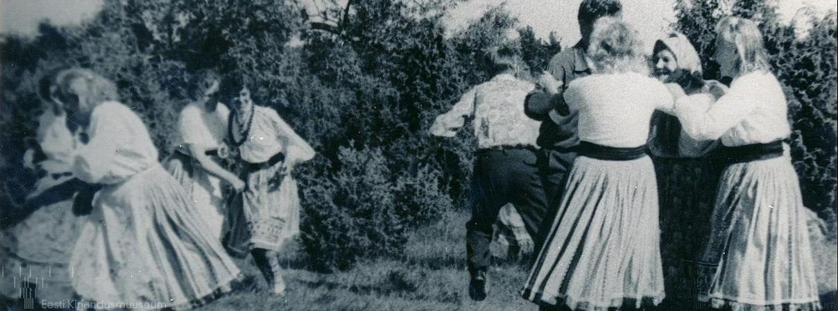 Vanal fotol on näha rahvariietes tantsivad rõõmsad inimesed ja tantsu rõõmsalt pealt vaatavad naised. Taustal on kadakad.