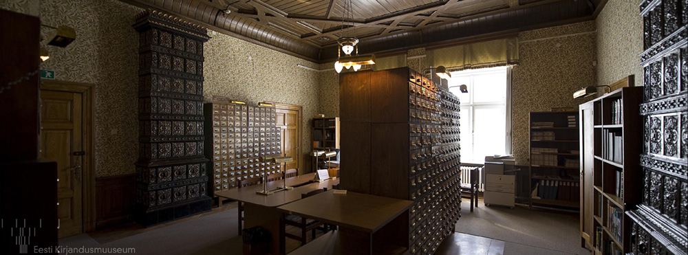 Arhiivraamatukogu kataloogisaal Fotol on Arhiivraamatukogu kartoteegikappidega puhas ja korras Foto on pruunikates toonides. Aknast paistab sisse ere päikesevalgus.